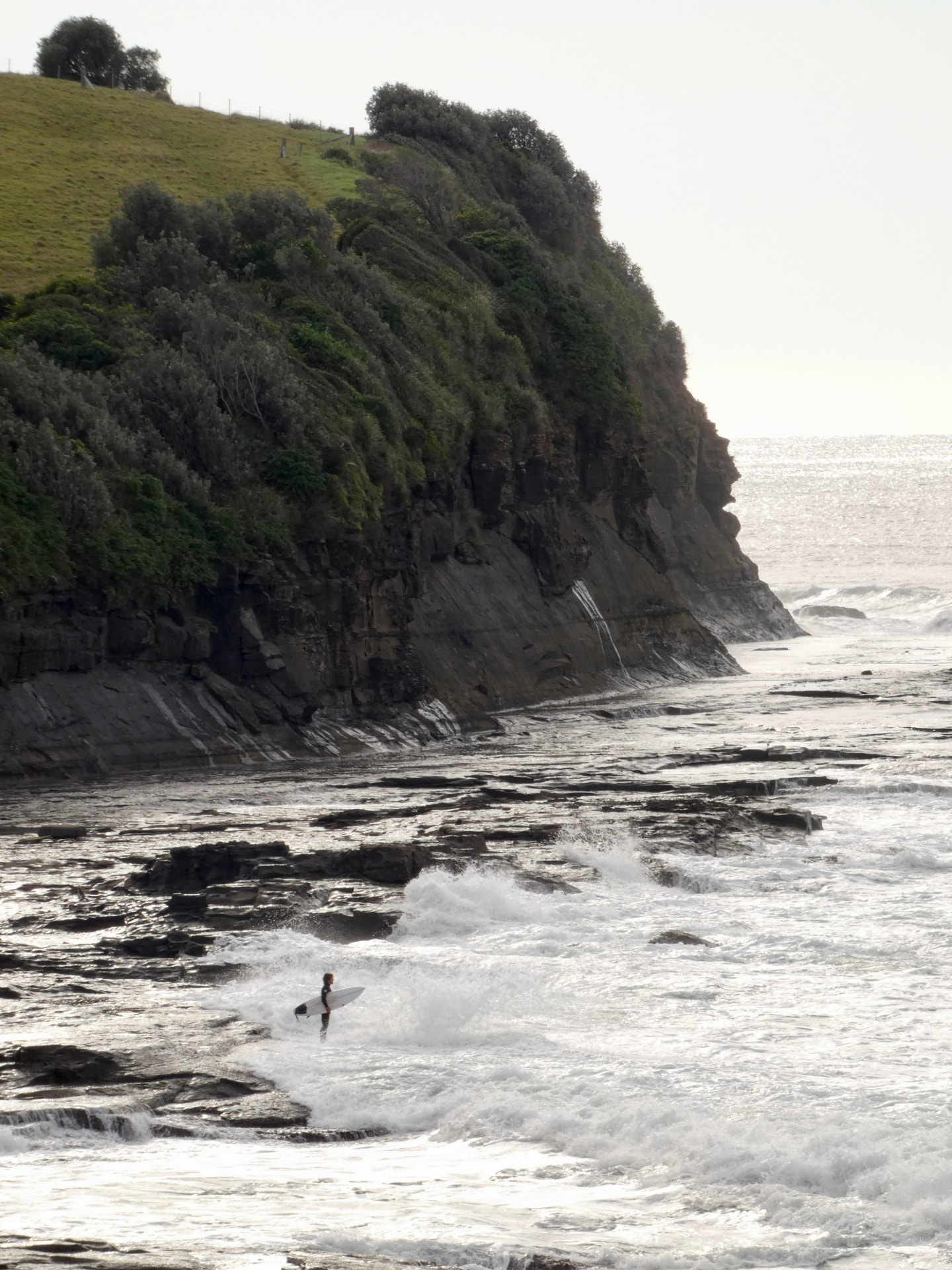 Lone surfer walking out below a green headland